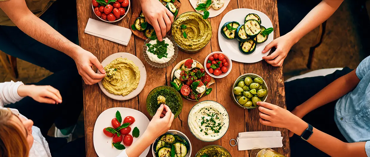 Top-down view of a wooden table with several hands reaching for antipasti dishes, such as grilled zucchini, dips, cherry tomatoes, and green olives.