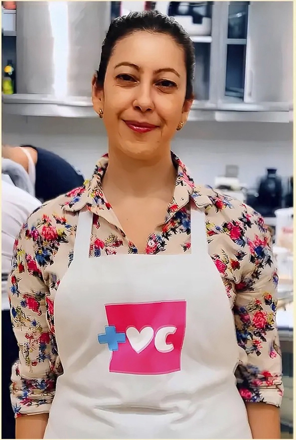 Grazie Canella wearing a floral shirt and apron with the Mais Você logo, smiling in a professional kitchen.