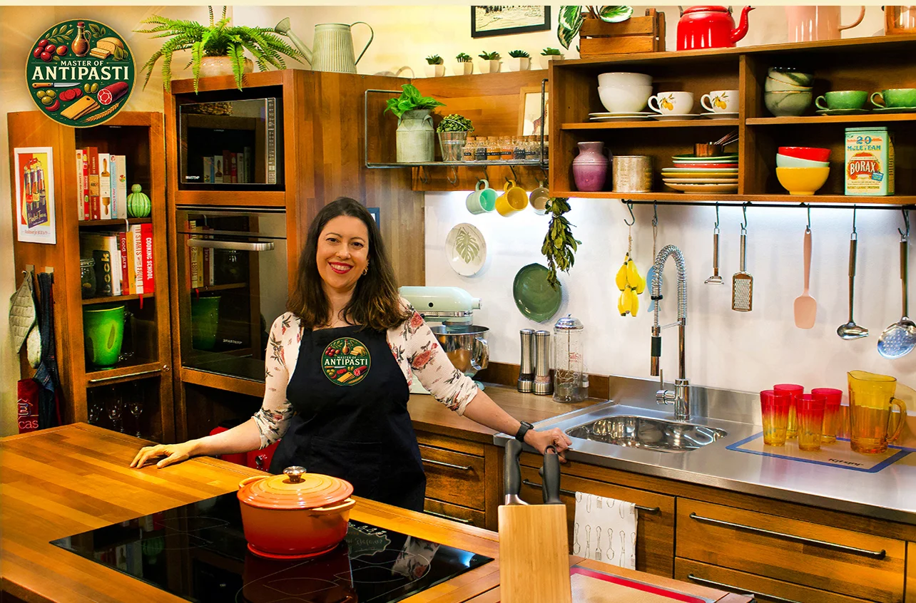 Grazie Canella standing in her colorful, well-equipped kitchen with a Master of Antipasti logo on the wall, smiling beside a counter with cookware.